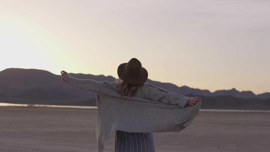 Women holds out her sweater as she walks forwards in slow motion wearing a hat towards the horizon of a sunset on a mountain range