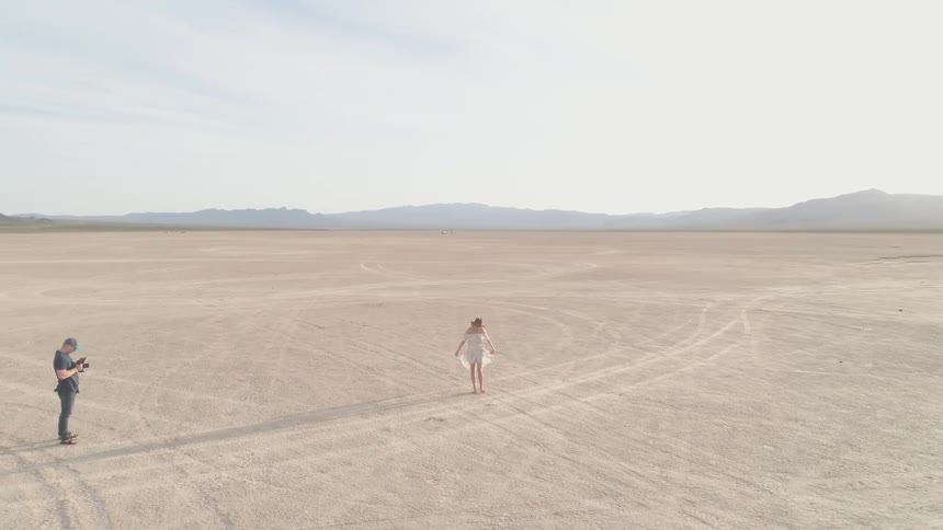 A woman stands in the middle of the desert with cracked dry soil in a white dress and a hat as the camera circles her and a cameraman can be seen with cars and trucks and video camera equipment