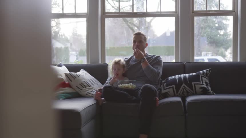Man sits on couch eating popcorn with small child