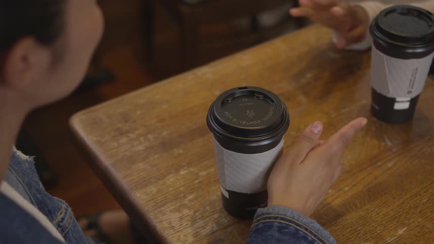 Two people have coffee at a small table with to go cups one is a woman with a jean jacket and they are discussing or conversing things with one another