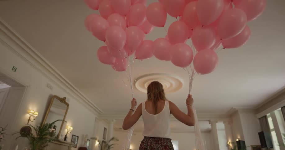 A woman carries a bundle of pink balloons in each of her hands across a house