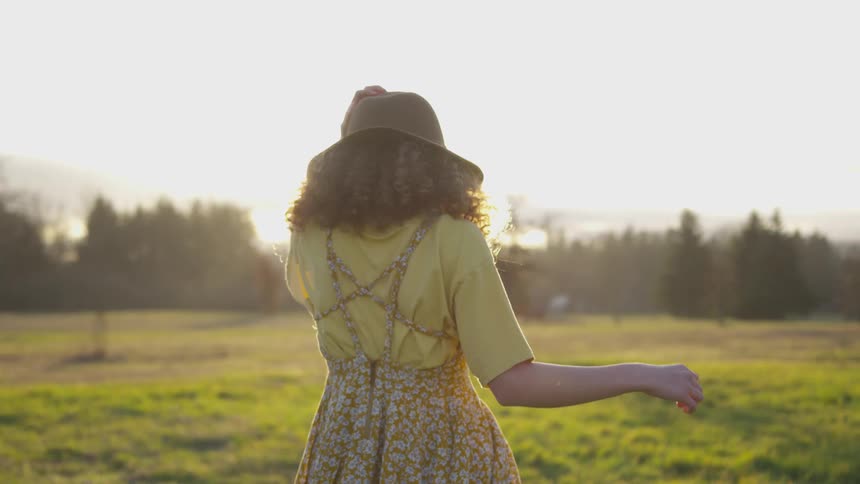 A woman touches her hat while walking in a sunny field