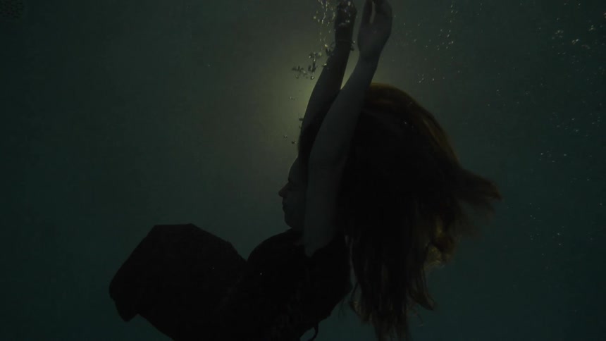 A woman sinks down into water as a light shines down from the surface