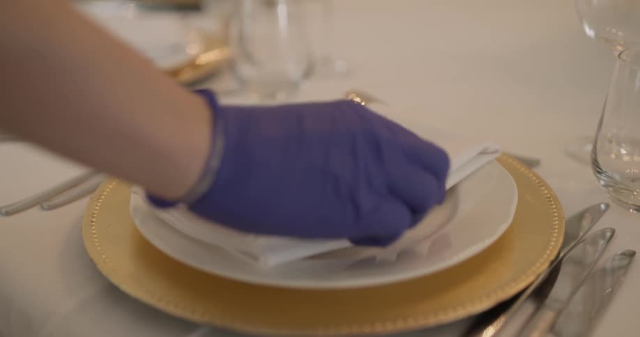 A gloved person placing cloth napkins onto a table setting