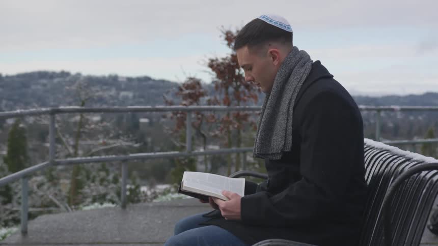 A man wearing a yamaka sits on a bench reading a book
