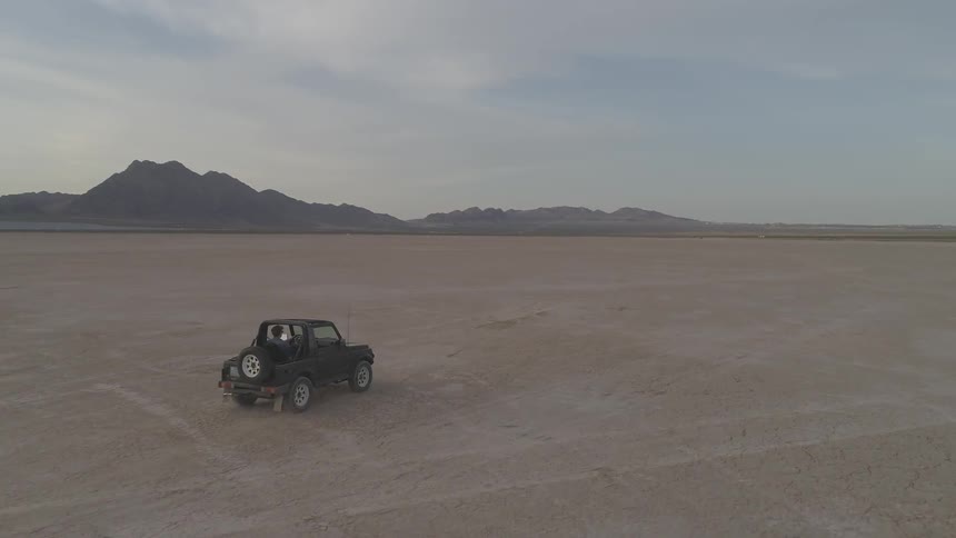 Jeep drives along wide empty plain past rocky hills