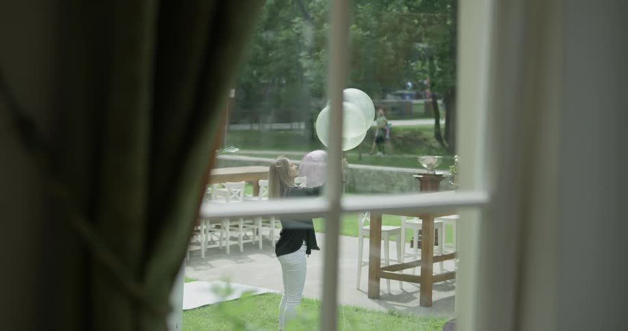 A woman with gray hair gathers some balloons from outside while standing in grass out in front of an establishment