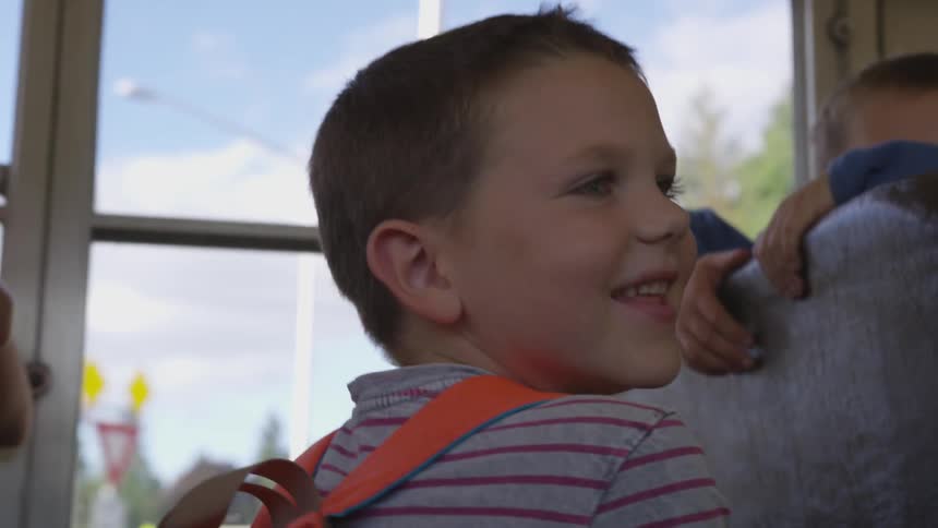 A boy wearing a backpack is looking out the bus window then turns around smiling