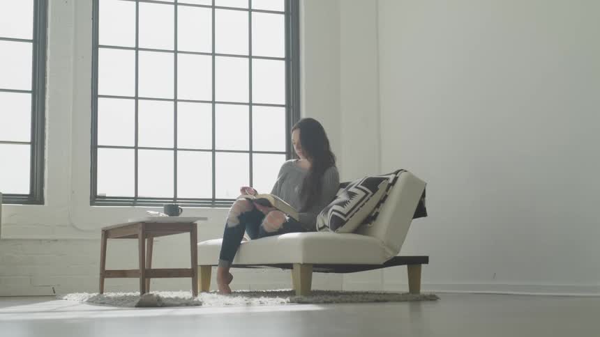 A dark-haired white female in ripped jeans sits on an ivory sleeper sofa reading a book in front of two large windows