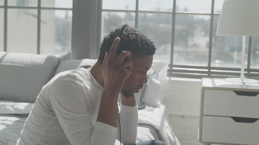 A black man in a bedroom puts on headphones