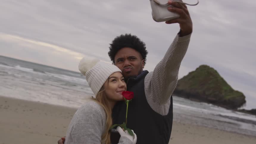 Man hugs girl who holds red rose and take a selfie on beach on a cloudy day