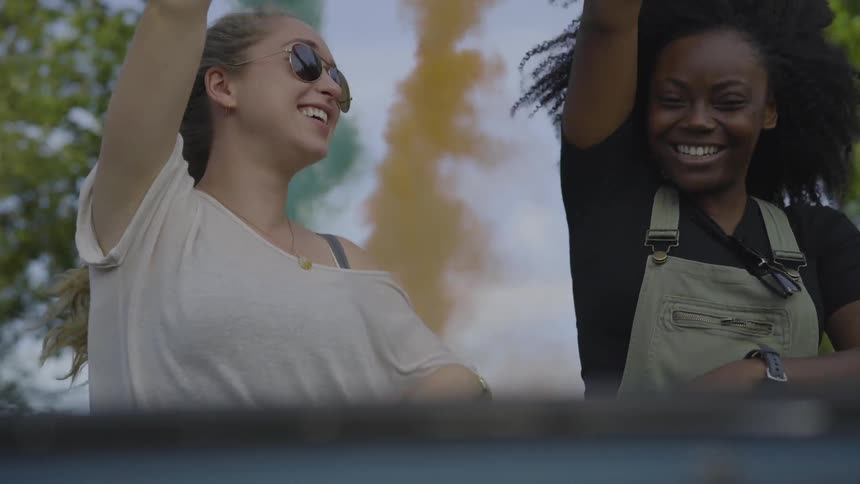Women wave blue and yellow smoke flares out of convertible