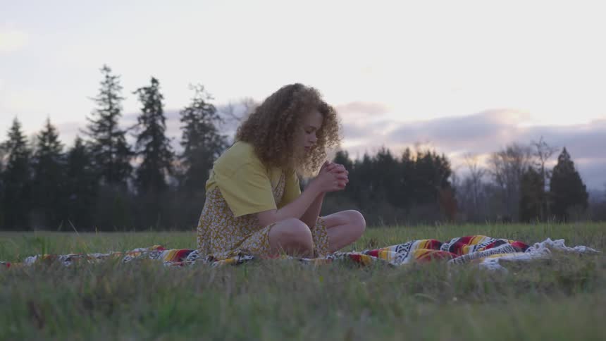 Woman prays while sitting on a blanket in the field