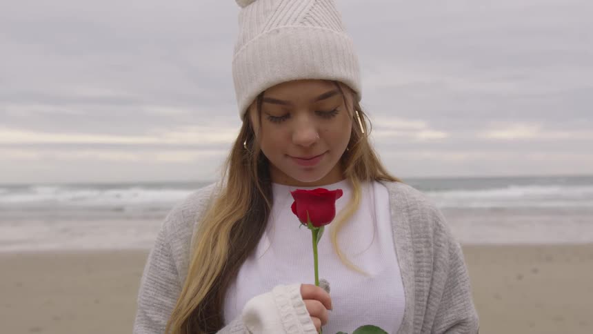 A woman stands at the beach holding a single red rose