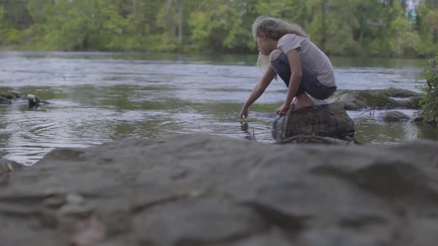 Young girl sits on rock in river tracing shapes in water with finger