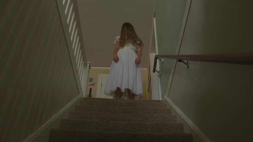 Creepy girl in white dress stands at the top of a staircase inside a house almost looks like a ghost in a dim lit room indoors