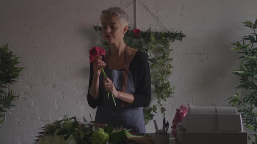 A young white short haired women smells flowers she is preparing on a desk in a dim lit room