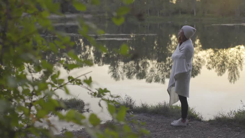 A woman stands near still water looking into the sky