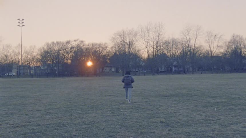 A child wearing a backpack walks across a grassy field towards homes at sunset