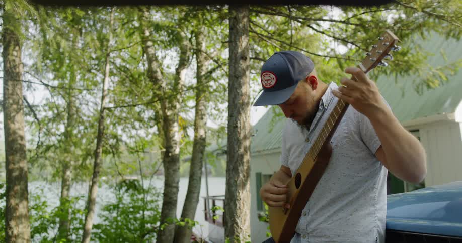 A man leaning on a car outside while playing a guitar
