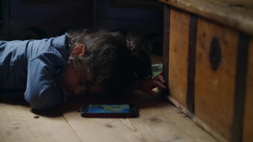 A toddler rests on the floor while a phone displays a cartoon