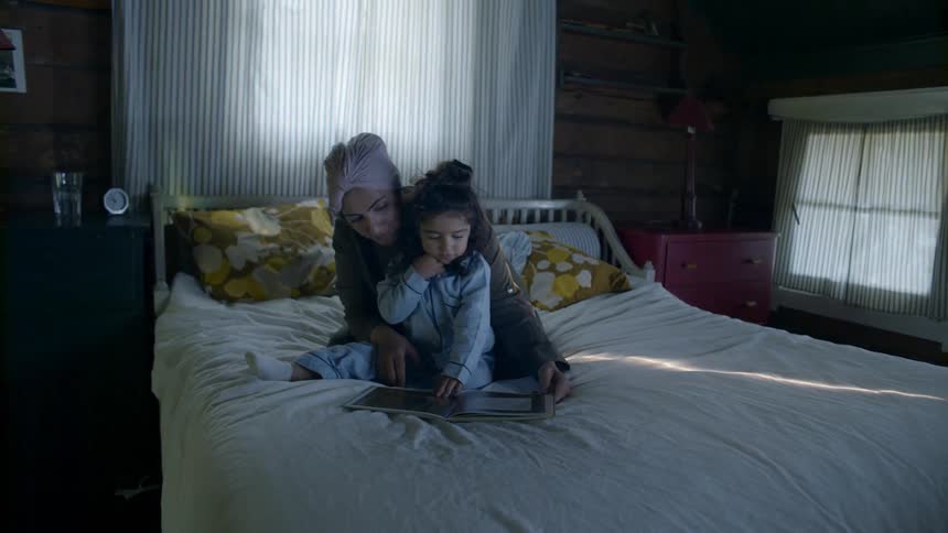 A woman and a little girl look at a tablet in a bed