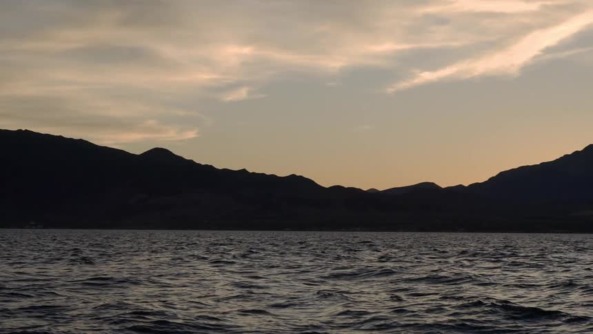 Slightly wavy lake is shown with clouds and hills in background at sunset