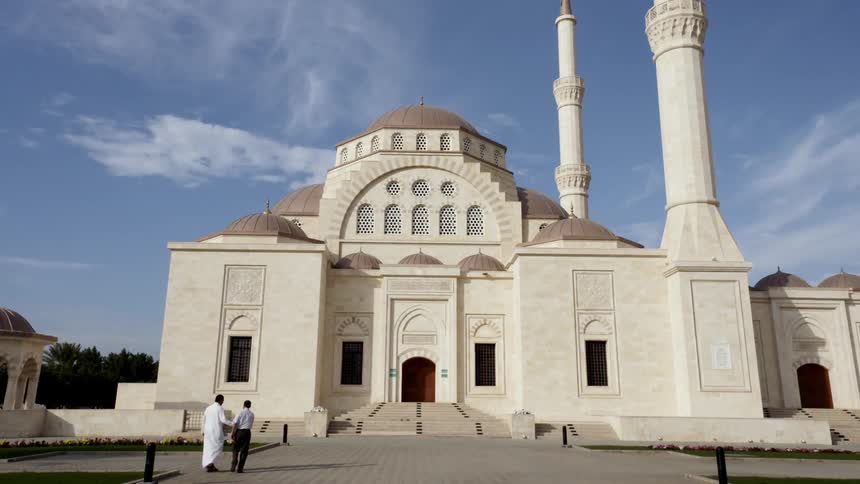 A beautiful mosque stands under a blue sky with wispy clouds as two men walk towards the entrance