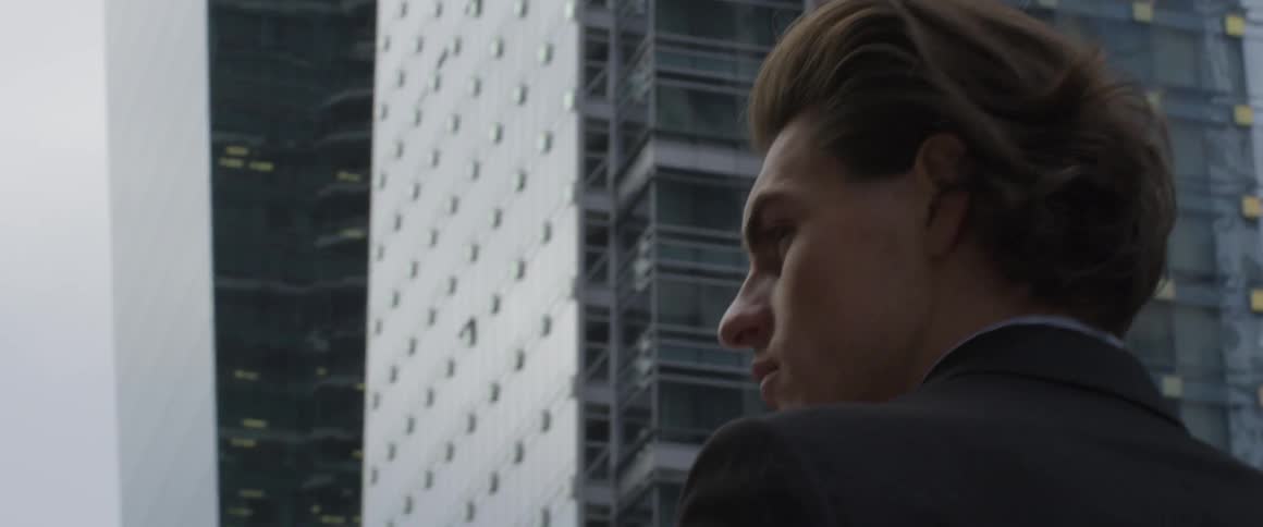 A man with swept back hair turns his head from the tall buildings in front of him