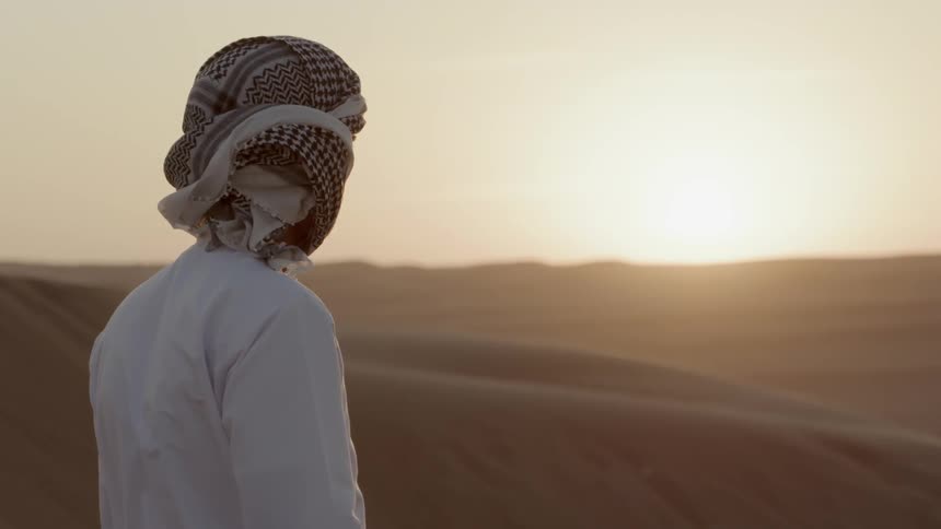 A man wearing traditional middle eastern garb stands in the desert looking at the sunset