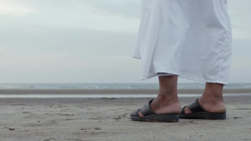 A person with sandals and a white gown stands on the beach