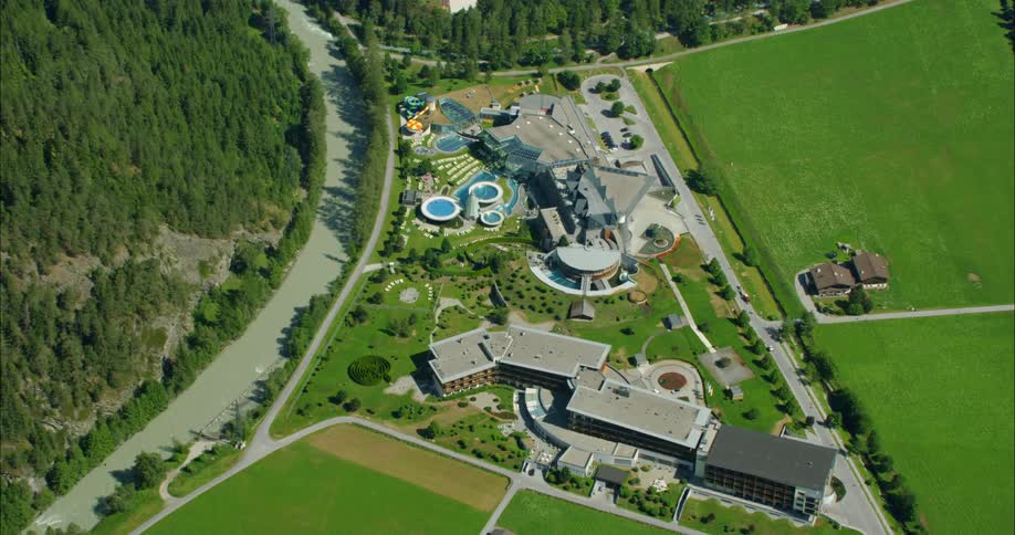 Two building complexes sit beside a large green field on one side and a forest on the other