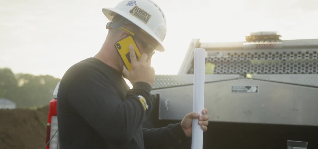 Man on Phone and Holding Blueprints at a Construction Site