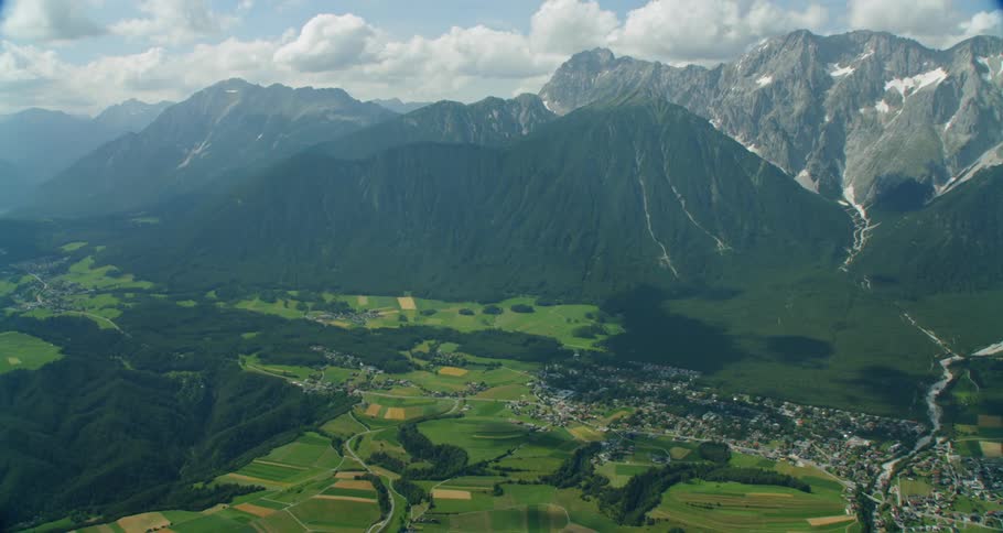 Green fields lead to large mountains covered in trees under puffy clouds
