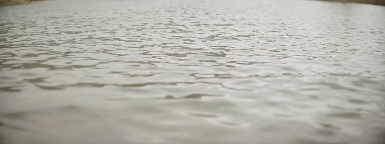 Rain falls into dirty lake as  one single man drifts blurredly