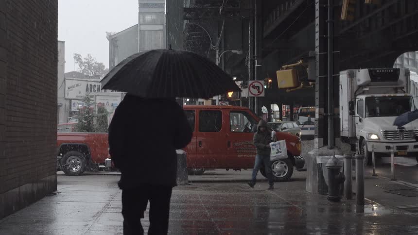 People walking in an urban area, some holding umbrellas over their heads, as it snows