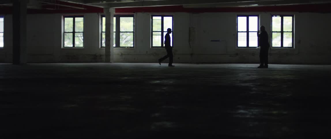 One man approaches another man in a window-lit empty building