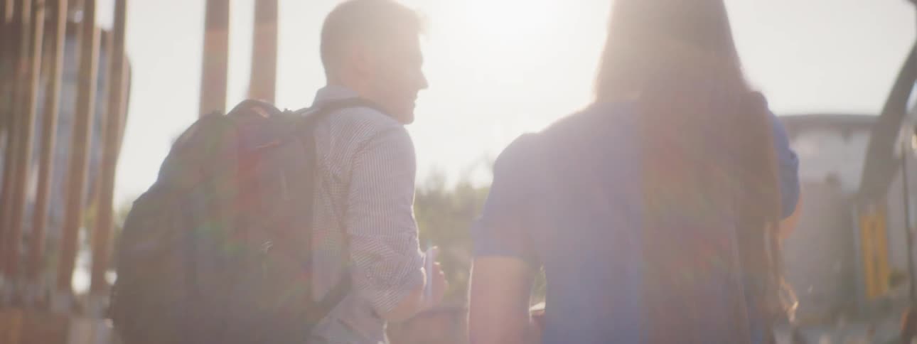 A man wearing a backpack and a woman carrying a bag walk away from a building and talk