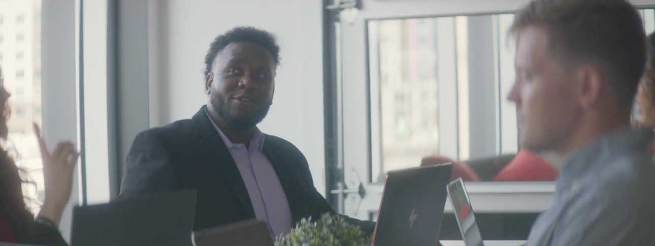 Man sitting at table with coworkers talks while sitting in front of laptop