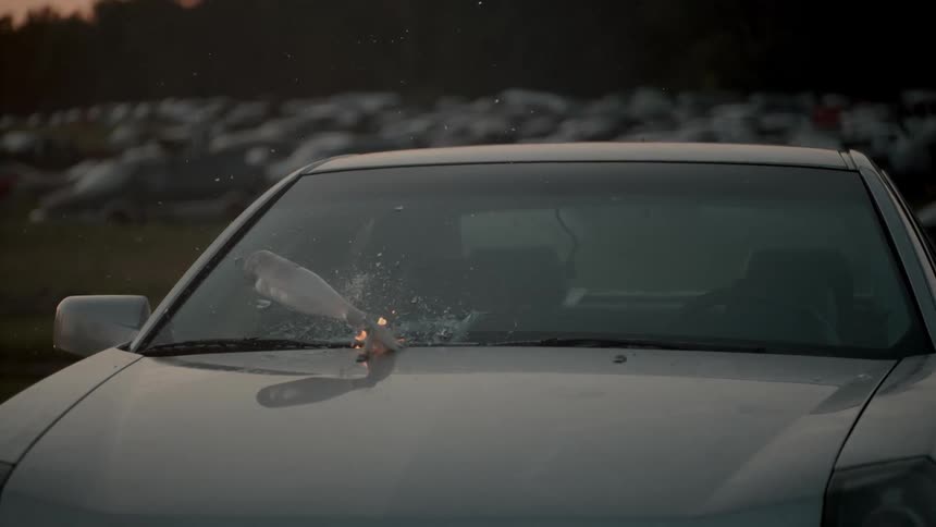 A flaming molotov cocktail lands on a car's hood then slides down the hood