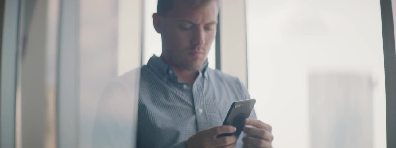 A man standing by a window types on his phone
