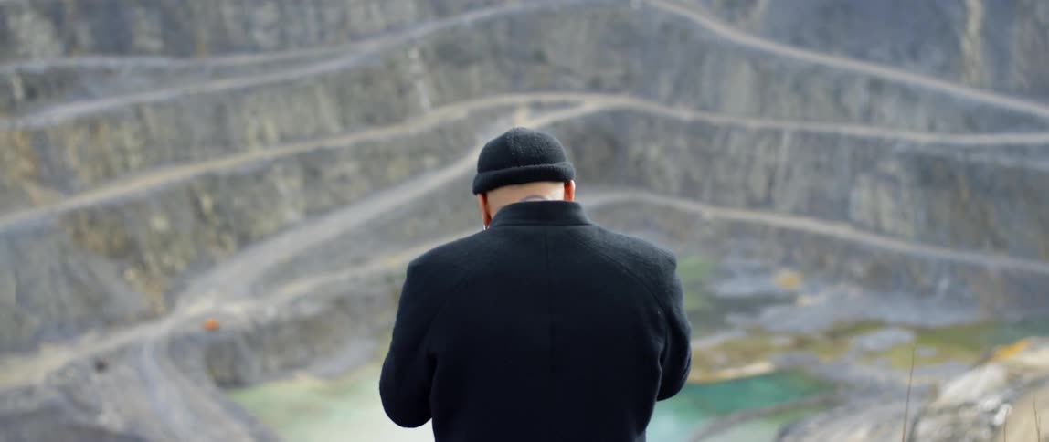 Man in hat  stands looking down onto road twisting up the side of a mountain during the day
