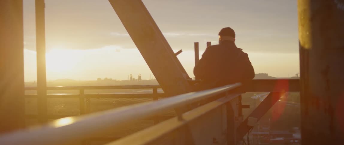 Man stands at rail near metals beams looking at city in distance near sunset