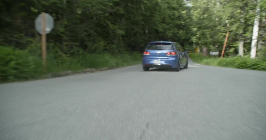 Blue car drives through an intersection and along a road bordered by thick woods