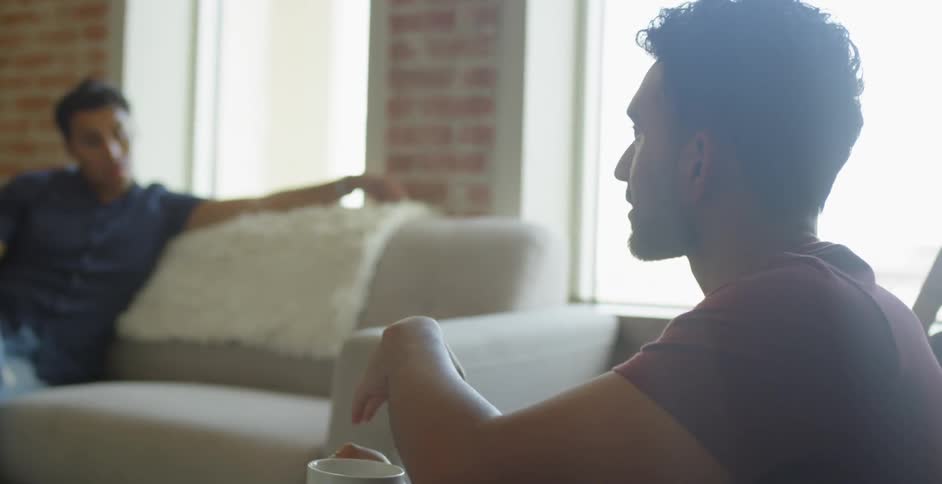 A man holding a cup talks with another man sitting on a couch