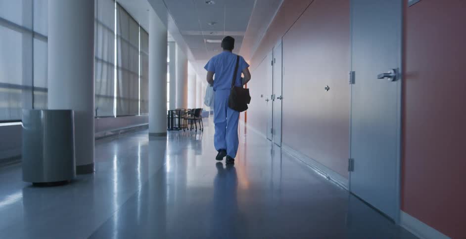 Man in scrubs carrying a white coat walks down a corridor near a wall of windows