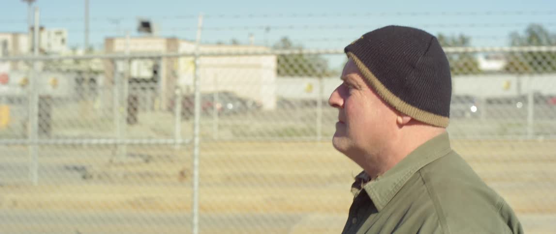 A man with a cap walks along a fence in town at day