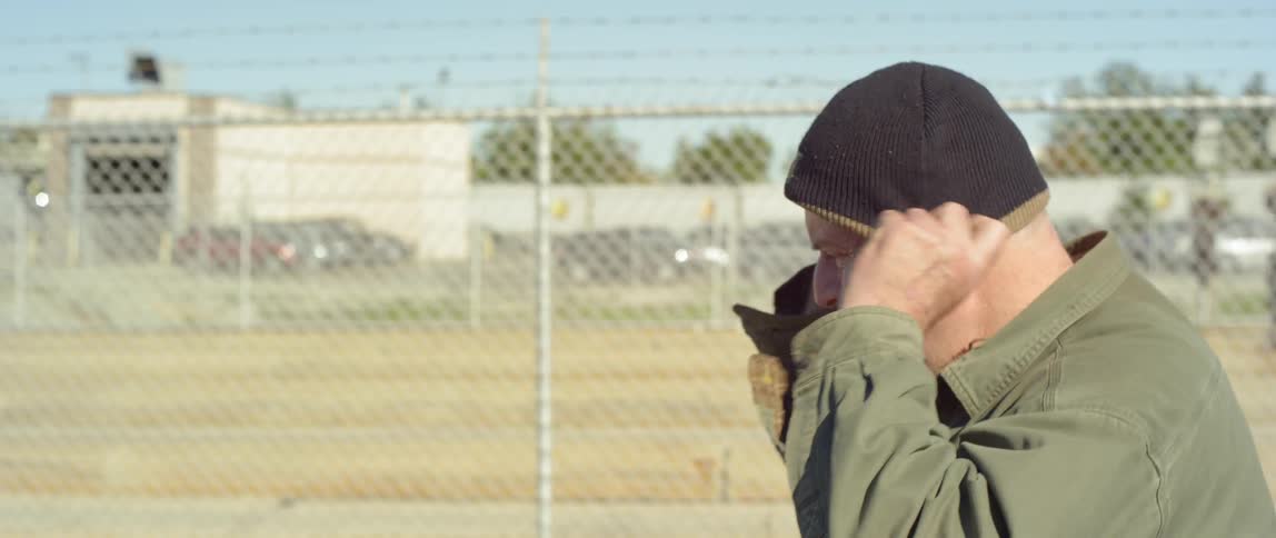 A man puts on a winter hat while walking next to a chain link fence