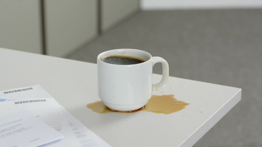 A man slams a white mug on a table and spills coffee next to some documents