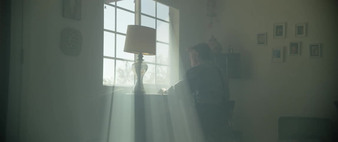 An elderly man sits at a desk in front of a sunny window
